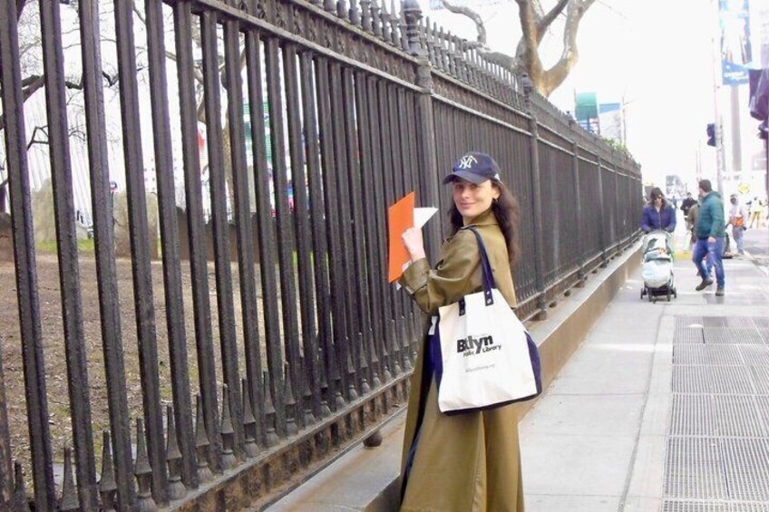 A person holding a shopping bag and a brochure, standing next to a metal fence. 3