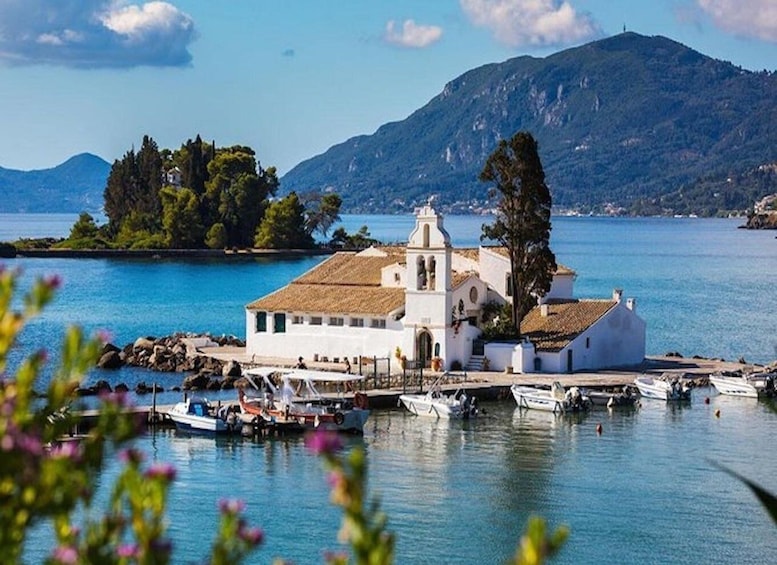A coastal church with a bell tower, surrounded by calm waters and boats, with a mountain in the background. 1