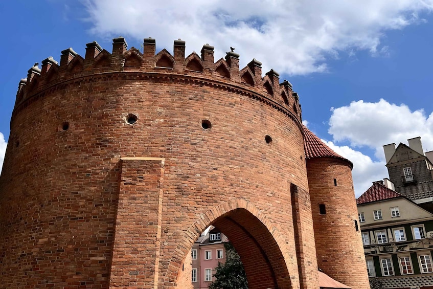 A brick tower with crenellations and an arched entrance, surrounded by buildings. 3