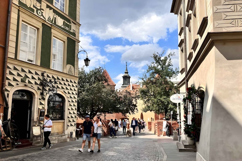 A cobblestone street lined with historic buildings, a church spire in the distance, and pedestrians walking. 1