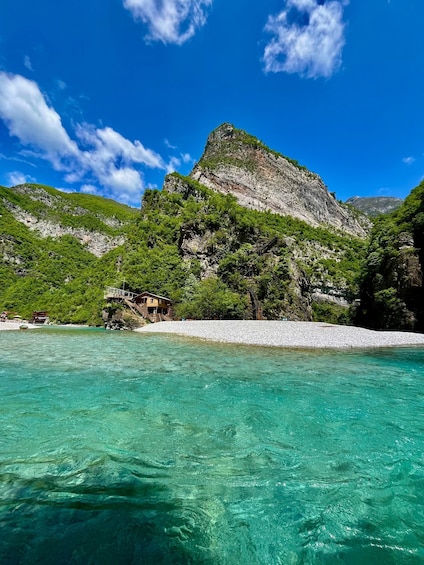 A coastal landscape with clear turquoise waters, a rocky hill with greenery, and a small building on the shore. 1
