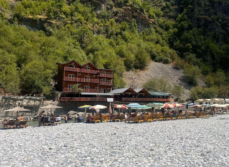 A beach with wooden lounge chairs and umbrellas, a wooden building with balconies, and a rocky hillside with trees. 5