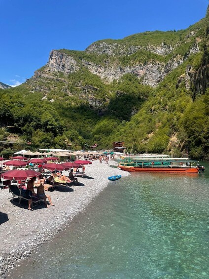 A pebble beach with umbrellas and people sunbathing, a boat, and mountains in the background. 4