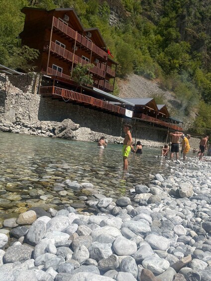 A clear river with people wading and standing in the shallow water, surrounded by large rocks. In the background, there is a wooden building with balconies and a red flag. 3