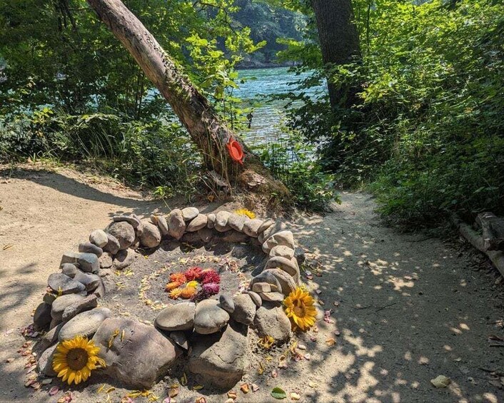 A circular stone arrangement with flowers and a red object on a dirt path by a lake. 3