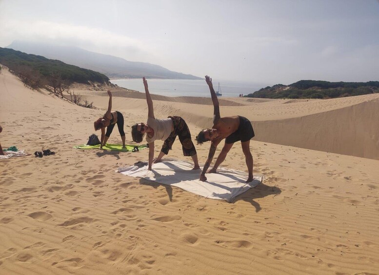 Three people practicing yoga on a sandy beach with a calm sea and hills in the background. 2