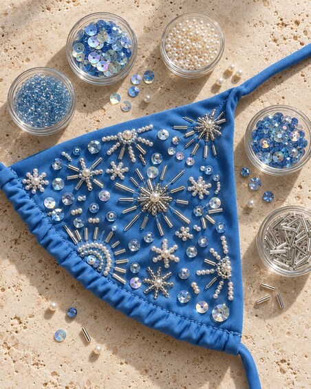Un tejido triangular azul decorado con lentejuelas, cuentas y elementos metálicos sobre una superficie arenosa. 3