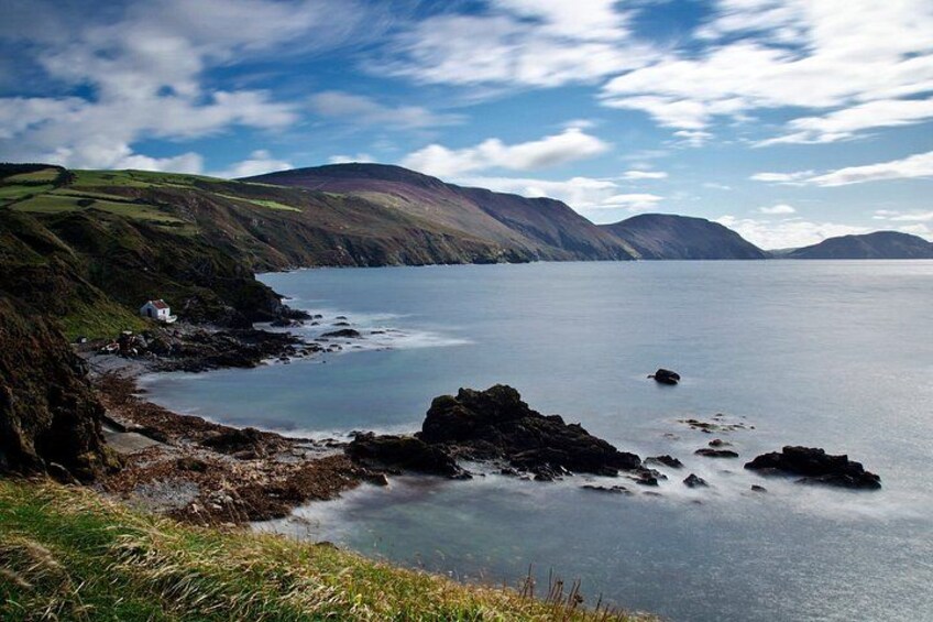 A coastal landscape with rocky shores, grassy hills, and a small house by the water. 1