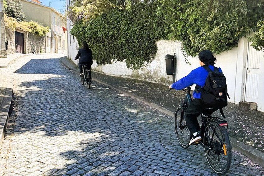 A cobblestone street with two cyclists, one in blue and one in black, riding away from the camera. 3