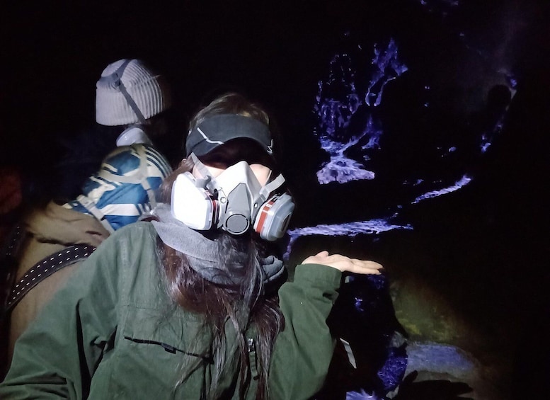 A person wearing a gas mask and a headlamp, reaching out towards a cave interior with blue lighting. 2
