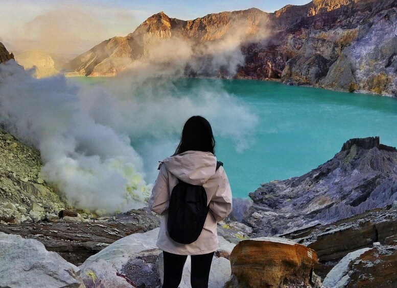 A person standing on rocky terrain overlooking a turquoise lake with steam rising from the water, surrounded by mountains. 1