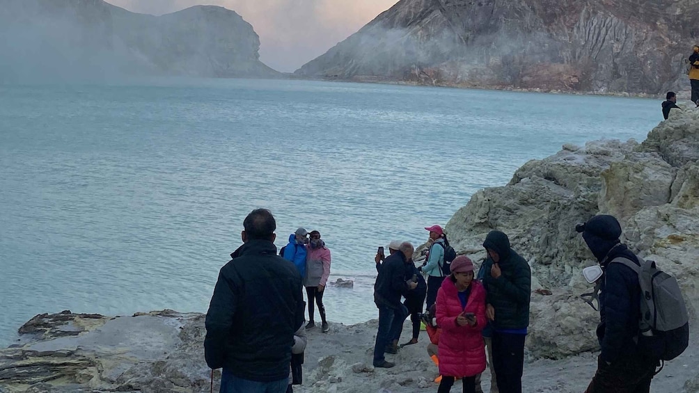 A group of people standing on a rocky shore by a calm lake with mountains in the background. 4