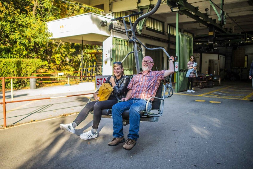 Two people on a stationary chair lift, with a building and greenery in the background. 1
