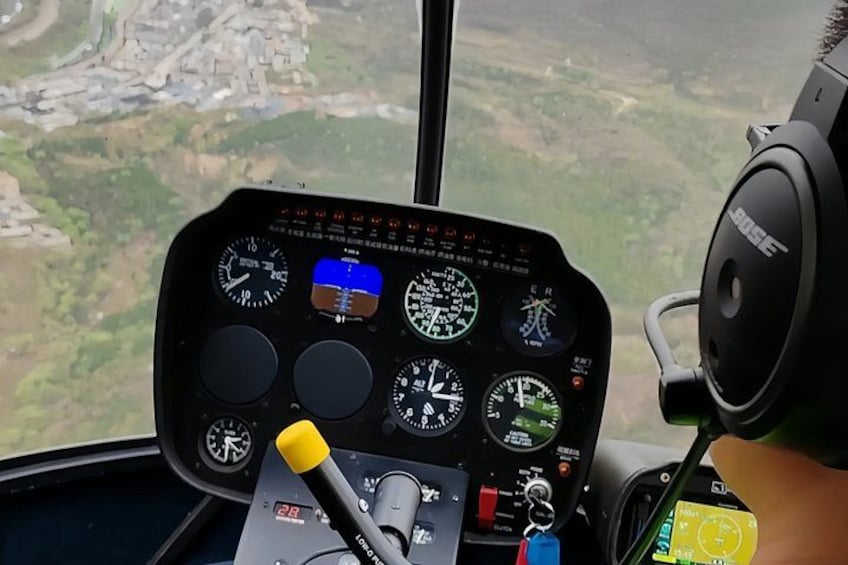 Helikoptercockpit med diverse instrumenter og betjening, udsigt over et landskab nedenfor, samt et sæt hovedtelefoner. 5