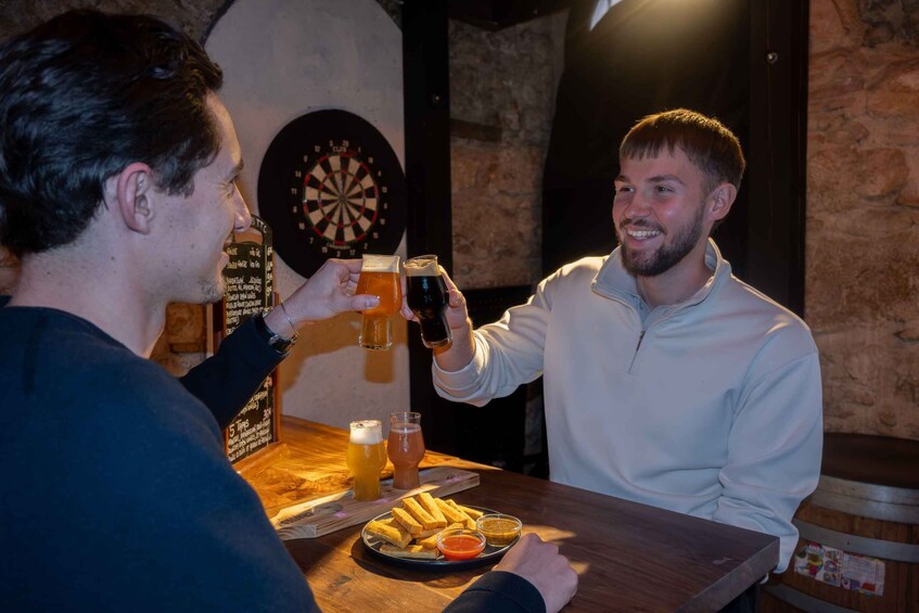 Two people toasting with beer glasses at a bar with a dartboard and a menu on the wall. 1