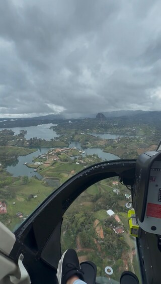 A helicopter cockpit with a view of a landscape featuring a lake, fields, and a mountain in the distance. 3