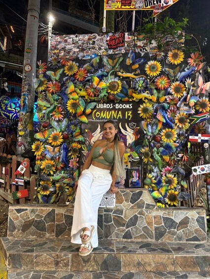 A person sitting in front of a colorful wall decorated with sunflowers and butterflies, with a sign that reads 'LIBROS - BOOKS COMUNA 13'. 2