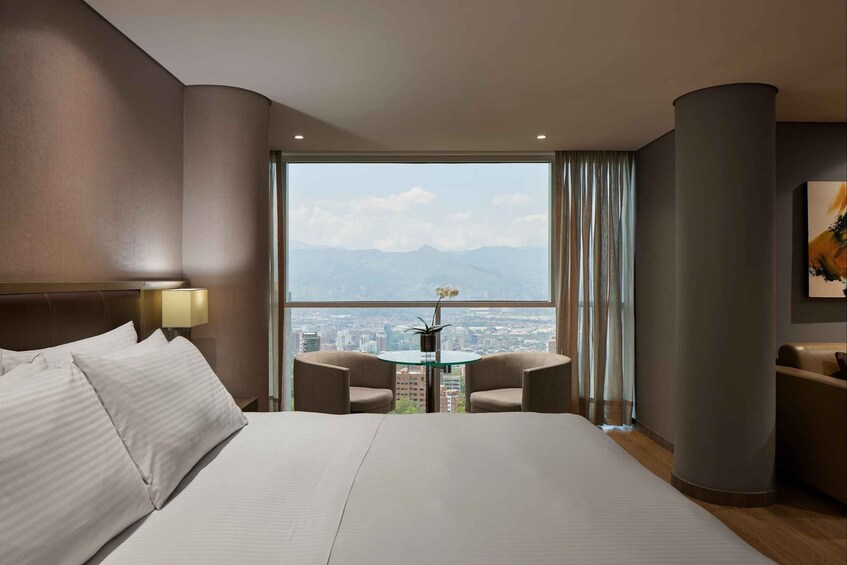 A hotel room with a large bed, two armchairs, a small table with a vase, and a cityscape view through the window. 5