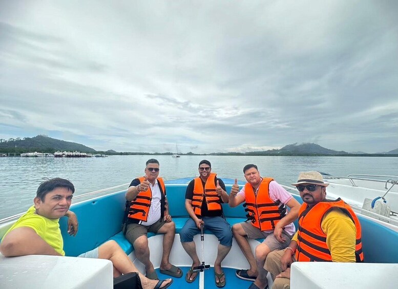 Five people on a boat wearing life jackets, with a lake and mountains in the background. 5