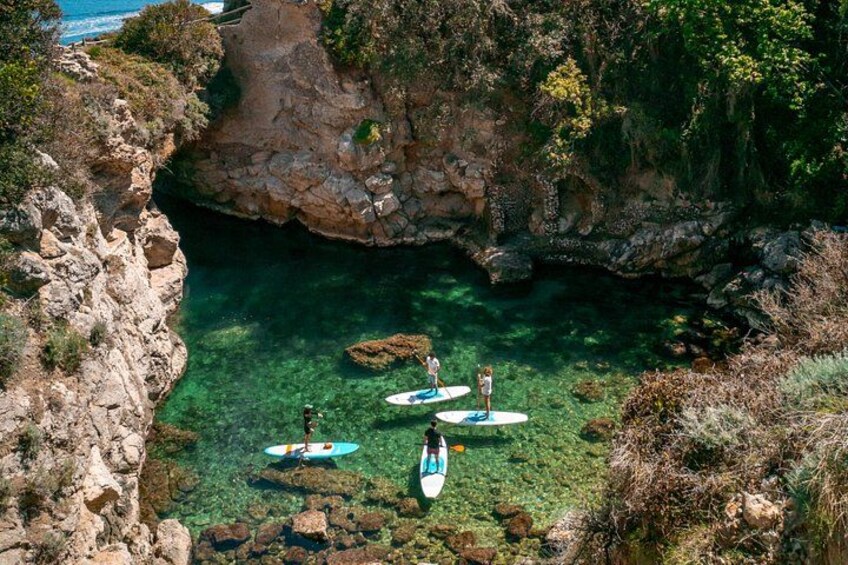 A coastal area with clear water, rocky cliffs, and people paddleboarding. 1