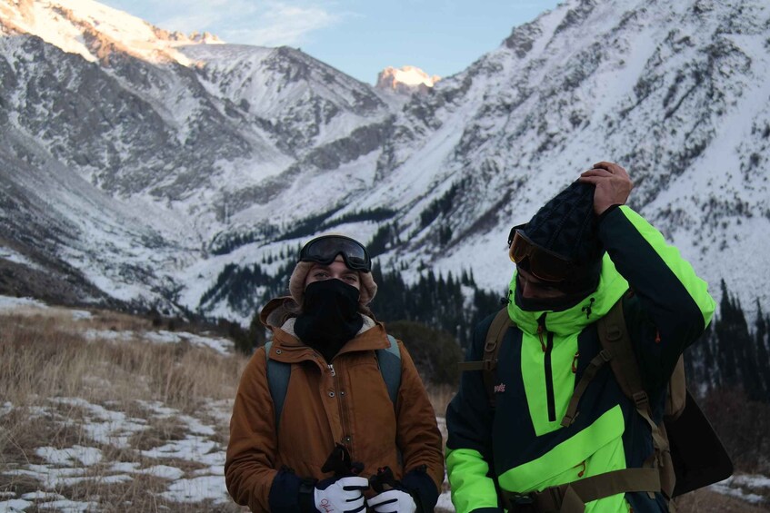 Two people in outdoor gear standing in a mountainous area with snow-covered peaks in the background. 3