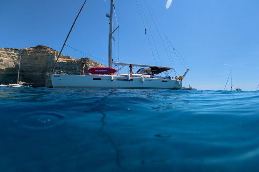 Ein weißes Segelboot mit einem roten Rettungsring auf ruhigem, blauem Wasser, mit Klippen im Hintergrund. 3