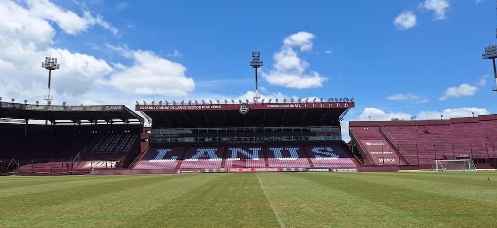 A stadium with 'LANUS' branding, empty stands, and a clear blue sky. 1