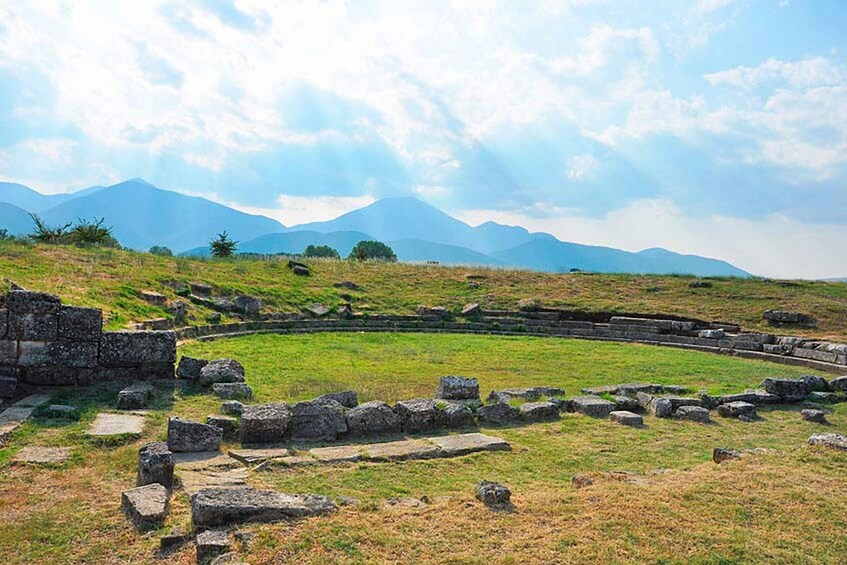 Ancient ruins with stone walls and a circular structure, set against a backdrop of mountains and a partly cloudy sky. 4