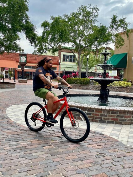 A person riding a red bicycle in a public square with a fountain and brick pavement. 5
