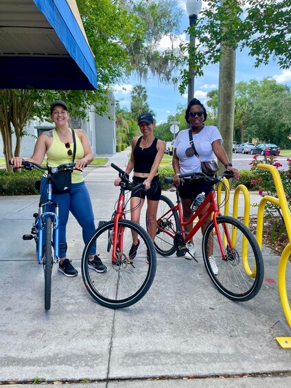 Three people standing next to bicycles on a sidewalk with trees and parked cars in the background. 3
