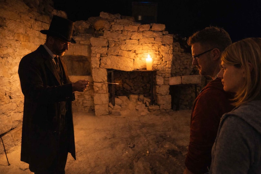 A man in a hat and coat standing by a stone fireplace with a lit candle, and two people observing him. 5