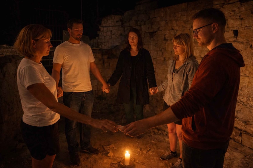 A group of people holding hands around a lit candle in a dimly lit stone-walled area. 3