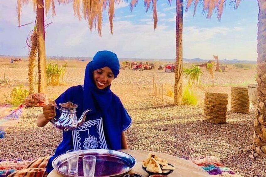 A person holding a teapot, seated at a table with a tray containing a glass and a plate of food, in a desert setting with palm trees and a clear sky. 4