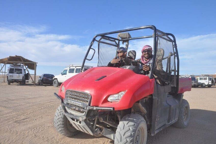 Un veicolo fuoristrada rosso con tre passeggeri, parcheggiato in un’area desertica con altri veicoli sullo sfondo. 2