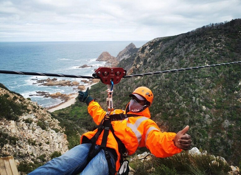 A person in an orange safety jacket and helmet, suspended by a zip line over a coastal landscape with rocky cliffs and the sea in the background. 2