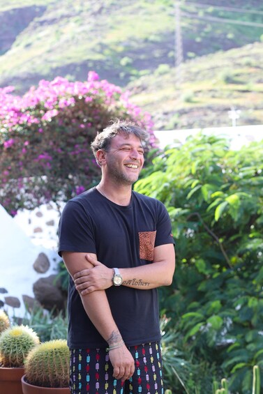 A person standing with arms crossed, wearing a dark t-shirt and colorful pants, with cacti and pink flowers in the background. 3