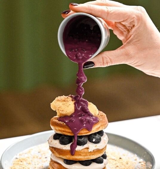 A stack of pancakes with blueberries and a hand pouring blueberry sauce. 3