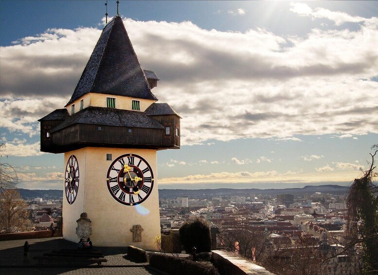 A clock tower with a pointed roof, Roman numerals, and a view of a cityscape in the background. 1