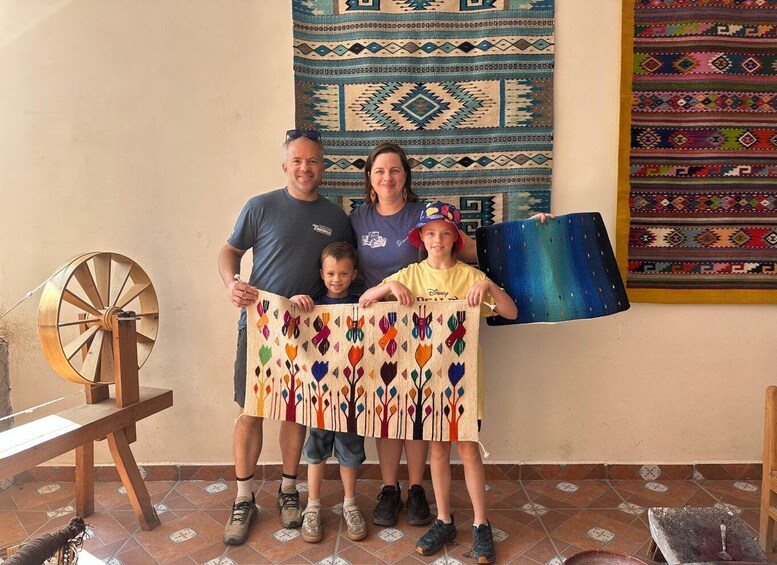 A family holding a colorful woven blanket, a spinning wheel, and a blue woven mat in a room with patterned rugs. 5