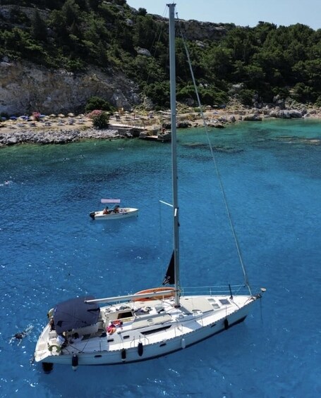 A sailboat anchored in clear blue waters with a rocky coastline and lush greenery in the background. 2