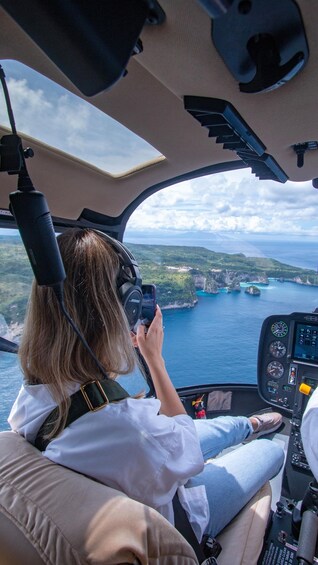 A person inside a helicopter cockpit, holding a camera, with a scenic view of a coastline and cliffs. 5