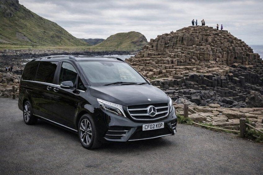 A black Mercedes-Benz minivan parked on a road with a rocky coastal landscape and a stone structure in the background. 1