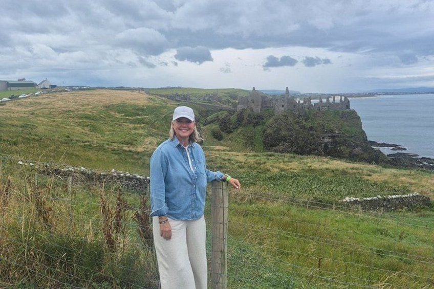 A person standing on a grassy hill with a stone wall, overlooking a coastal landscape with ruins and a body of water. 4