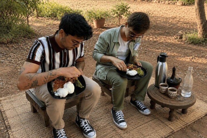 Two people sitting outdoors on a bench, eating from black plates with food and drinks. 2