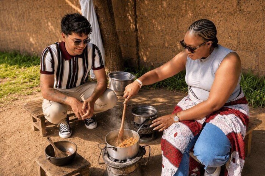 Two people cooking outdoors with a pot on a stove and a wooden stool. 1