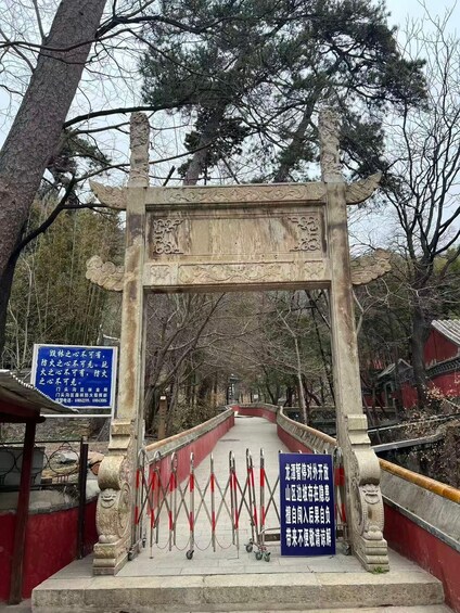 A traditional stone archway with intricate carvings, leading to a pathway flanked by red barriers and a blue informational sign. 3