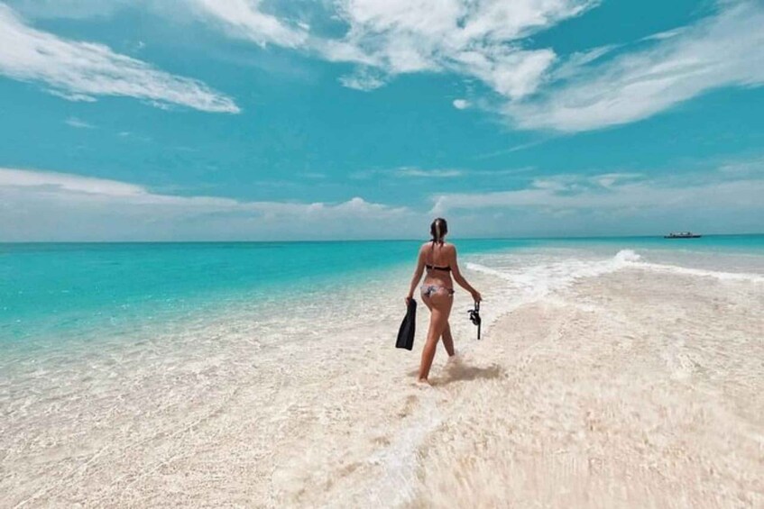 A person walking on a sandy beach with clear turquoise water and a boat in the distance. 2