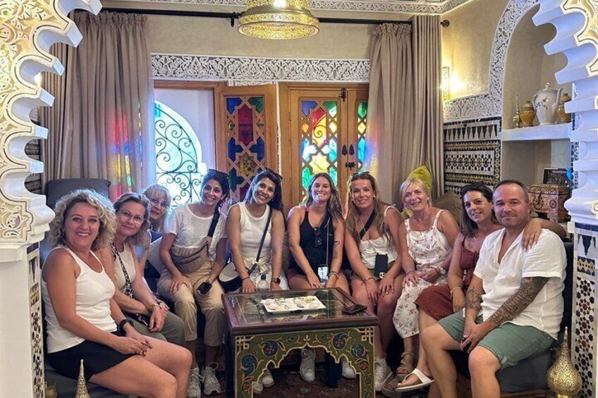 A group of people seated in a room with ornate decor, including a stained glass window and patterned furniture. 4
