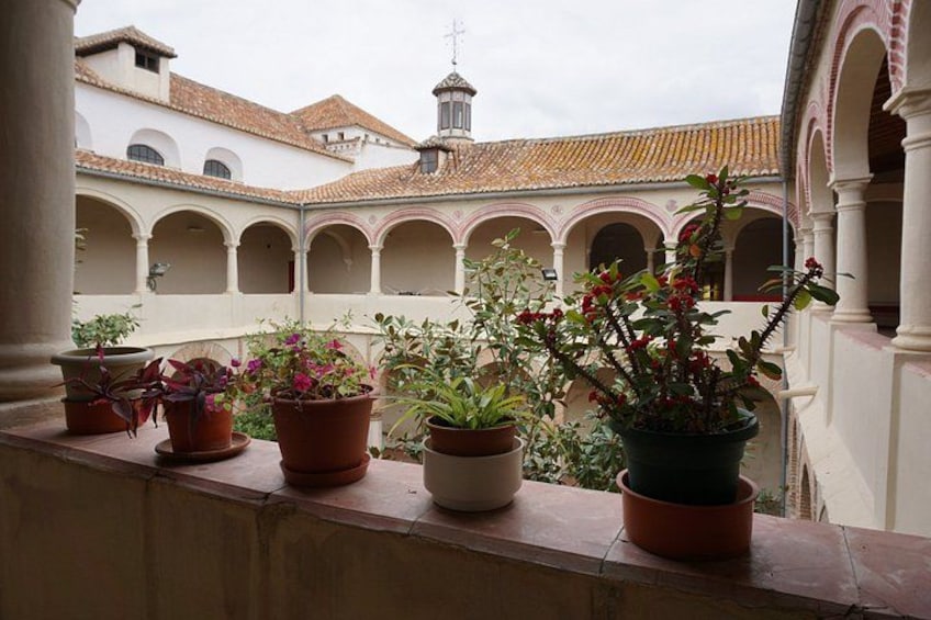 A balcony with potted plants overlooking a courtyard with arches and a building with a cross on top. 4