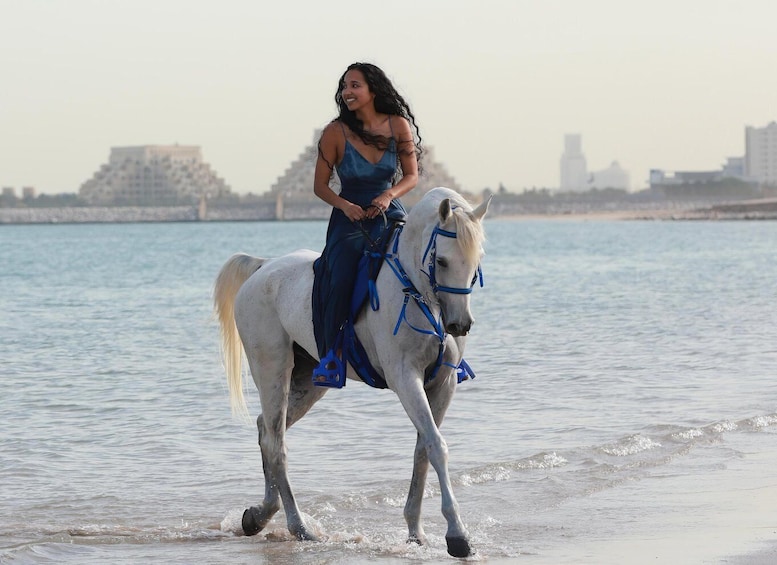 A person riding a white horse along a beach with water and buildings in the background. 1